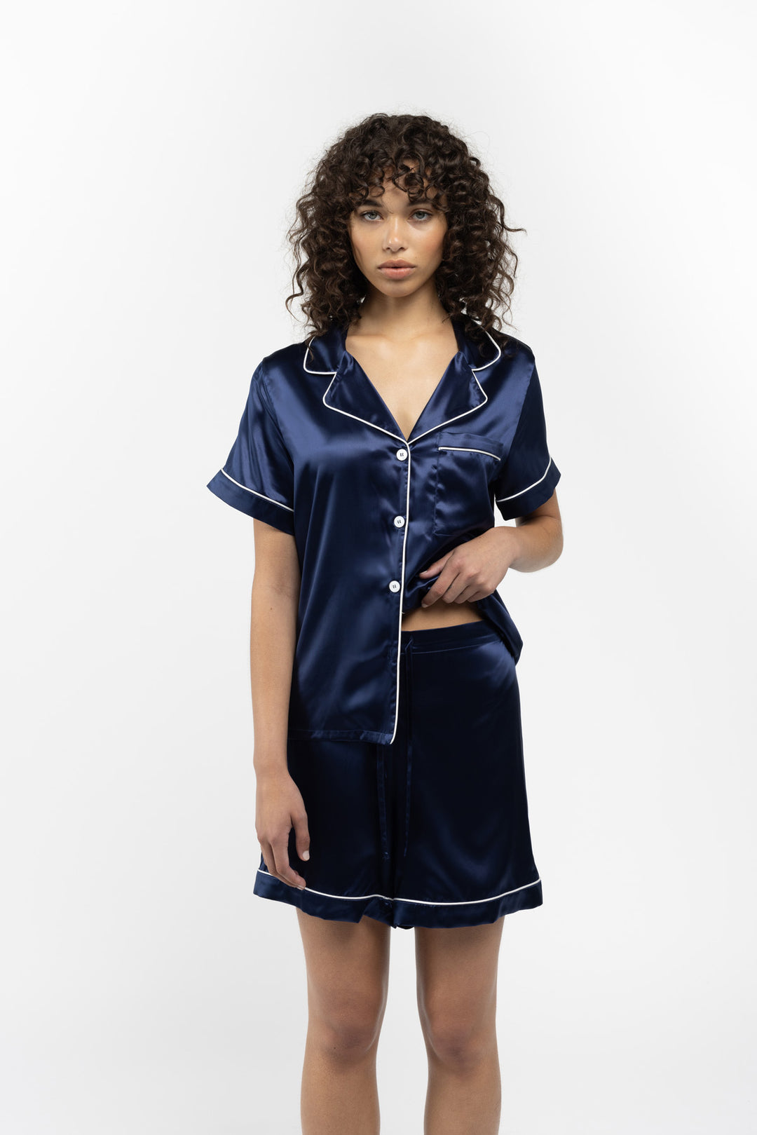 Model wearing navy silk short-sleeve button-up pajama top with white piping and matching shorts.