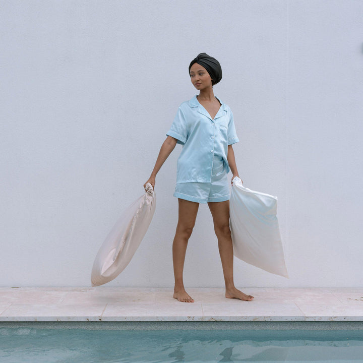 Person in light blue silk pajamas holding two pearl-white silk pillowcases beside pool and pale exterior wall.#color_pearl-white