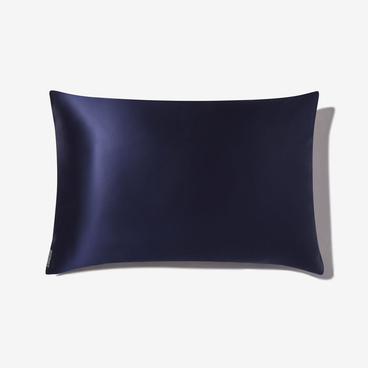 Navy blue silk pillowcase laid flat, rectangular with subtle sheen and small fabric tag at lower-left corner. - #color_navy-blue
