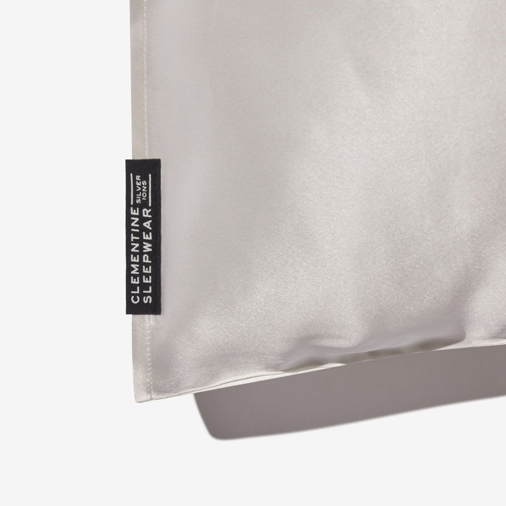 Pearl-white silk pillowcase corner with black tag reading CLEMENTINE SLEEPWEAR SILVER IONS - #color_pearl-white