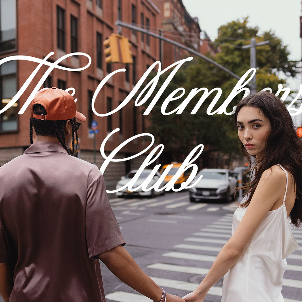 Couple holding hands crossing a city intersection with woman looking at camera; script text reads 'The Members Club'.