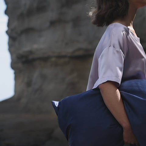 Woman holding a large navy-blue cushion, cropped at shoulder and hand with rocky cliff background. - #color_navy-blue