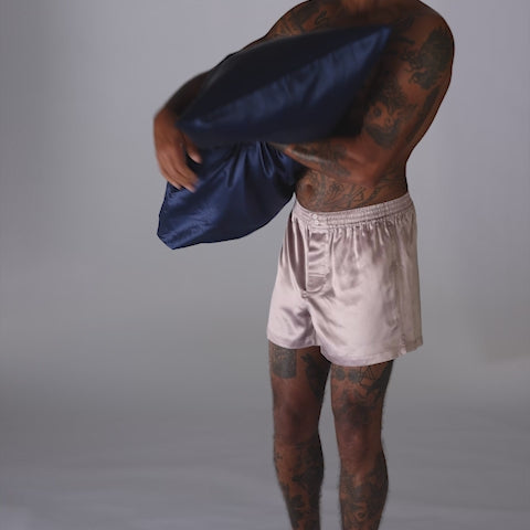 Tattooed torso and legs of a man wearing pale pink silk boxer shorts, holding a navy-blue silk pillowcase - #color_navy-blue