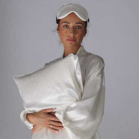 Person in a pearl-white silk robe holding a white pillow and wearing a sleeping mask. - #color_pearl-white