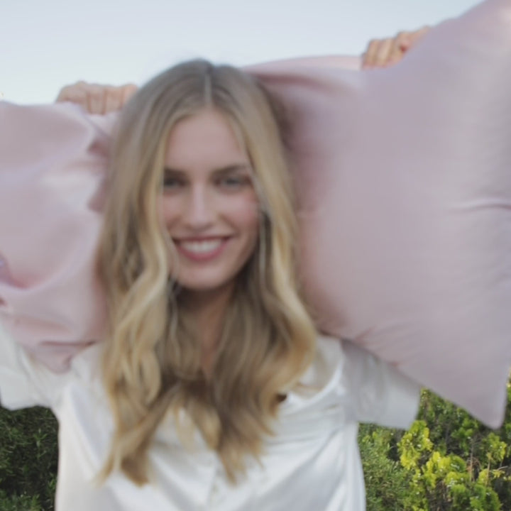 Smiling woman outdoors holding two pink silk pillowcases behind her head - outside with pink pillowcase in silk#color_pink-sherbert