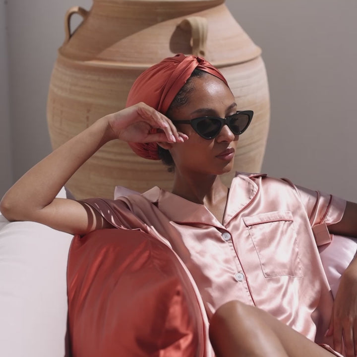 Reclining woman outdoors on sofa in pink silk pajamas, coral headwrap and black cat-eye sunglasses; terracotta urn behind - Lying outside with dusk pillowcase under head#color_dusk