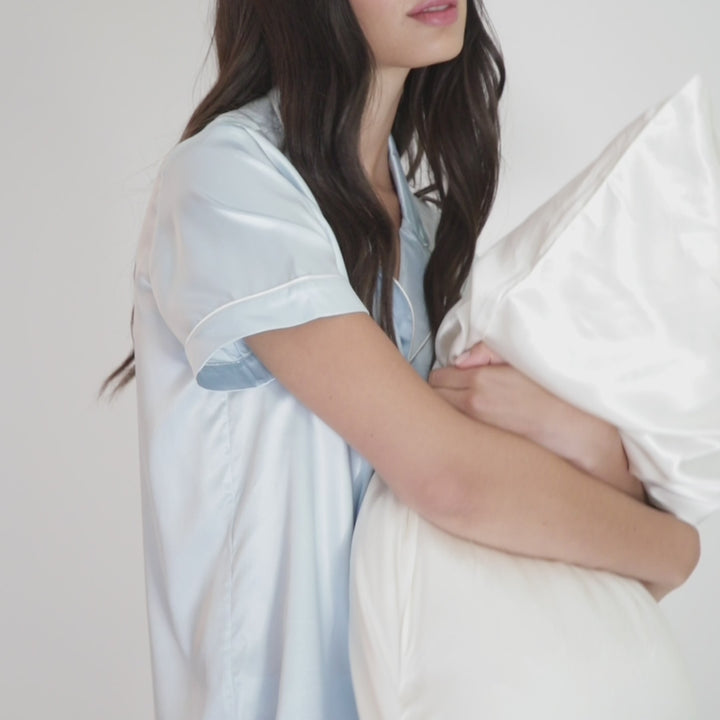 Person in light-blue silk pajamas hugging a pearl-white organic silk pillowcase.#color_pearl-white