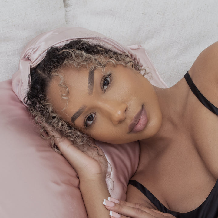 Close-up portrait: woman resting on pink silk pillow with curly hair, silk headscarf and French-manicured hand near her face - curly hair on pink pillowcase#color_pink-sherbert