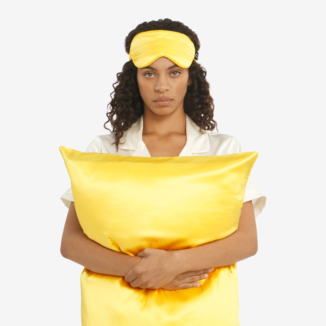 Woman in cream pajamas holding a lemon-yellow satin pillowcase, front view, wearing a matching yellow sleep mask.#color_lemon