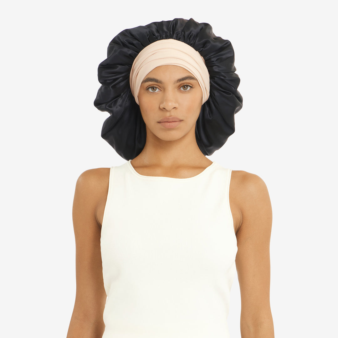 Front-facing portrait of woman wearing a large black satin bonnet over a beige headwrap and sleeveless white top.