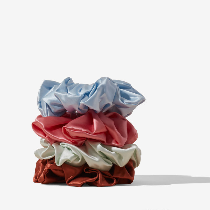 Stack of four silk hair scrunchies in pale blue, coral pink, mint, and rust on a neutral background.