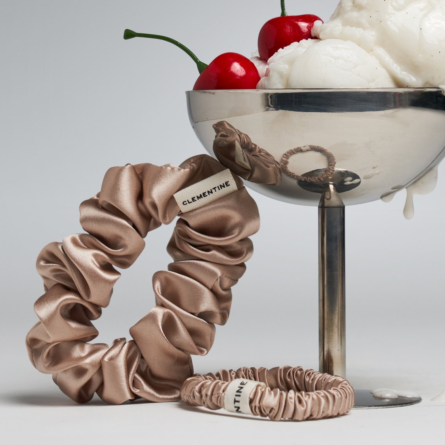 Two beige silk scrunchies labeled 'CLEMENTINE' next to a metal bowl with ice cream and cherries