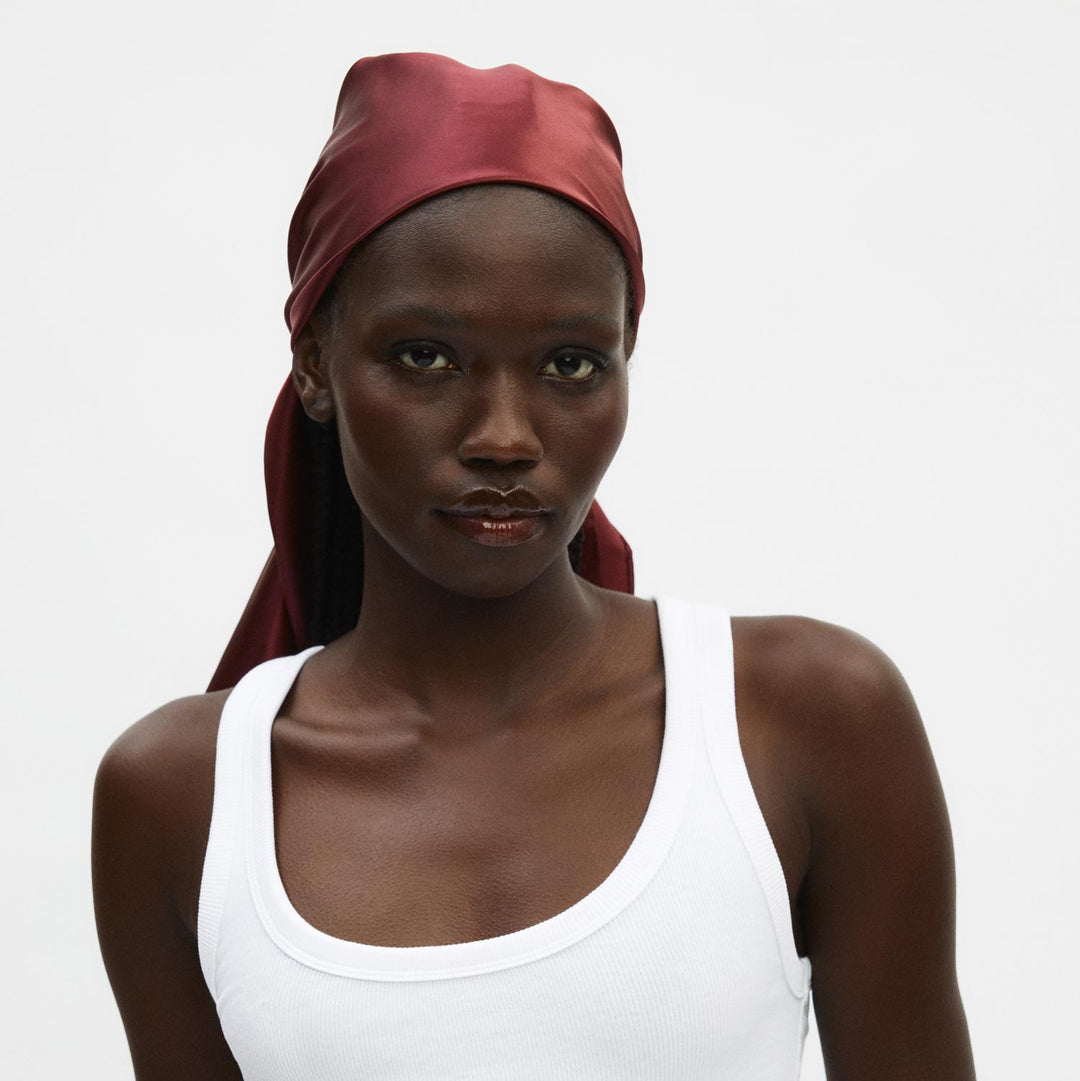 Model wearing burgundy silk sleep scarf tied at the back, shown chest-up in a white tank top.