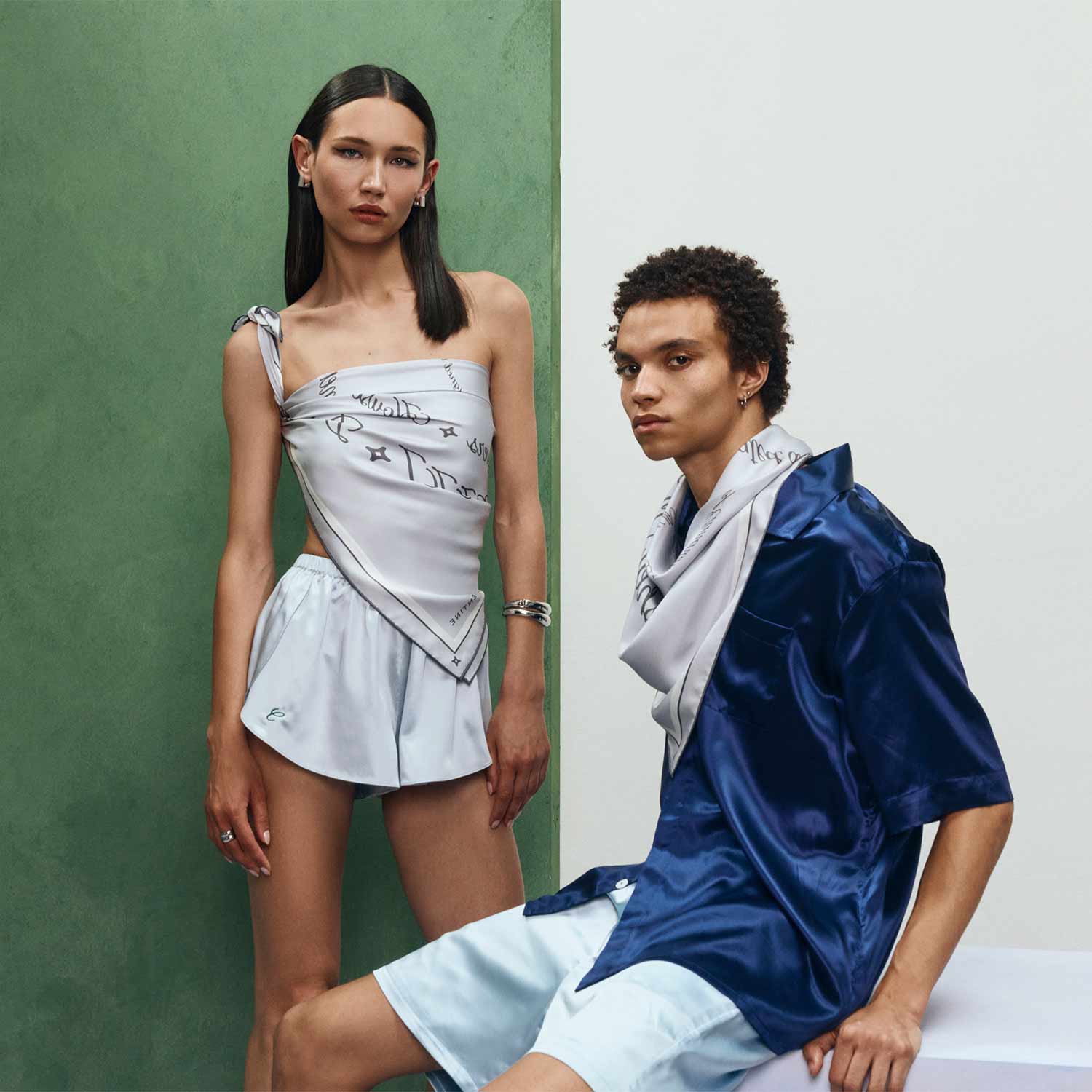 Two models pose, woman in light silver silk top and shorts, man seated in navy silk shirt and light blue shorts #color_silver-and-stone