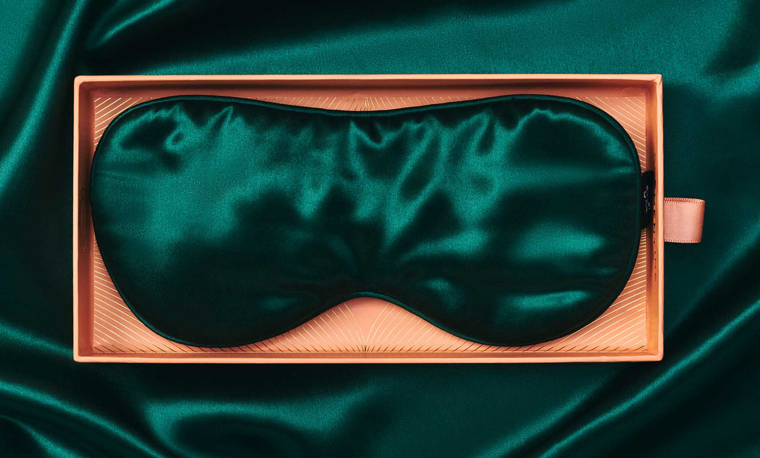 Top-down view of emerald silk sleep mask in a pink gift box, contoured padded shape with black trim on silk background