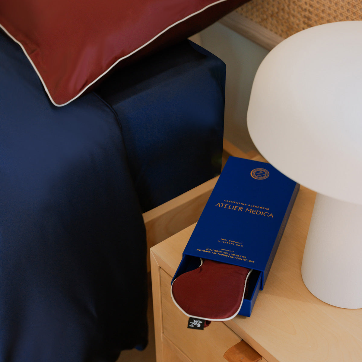 Close-up of navy silk fitted base sheet with Atelier Medica packaging and red silk eye mask on nightstand #color_navy-blue