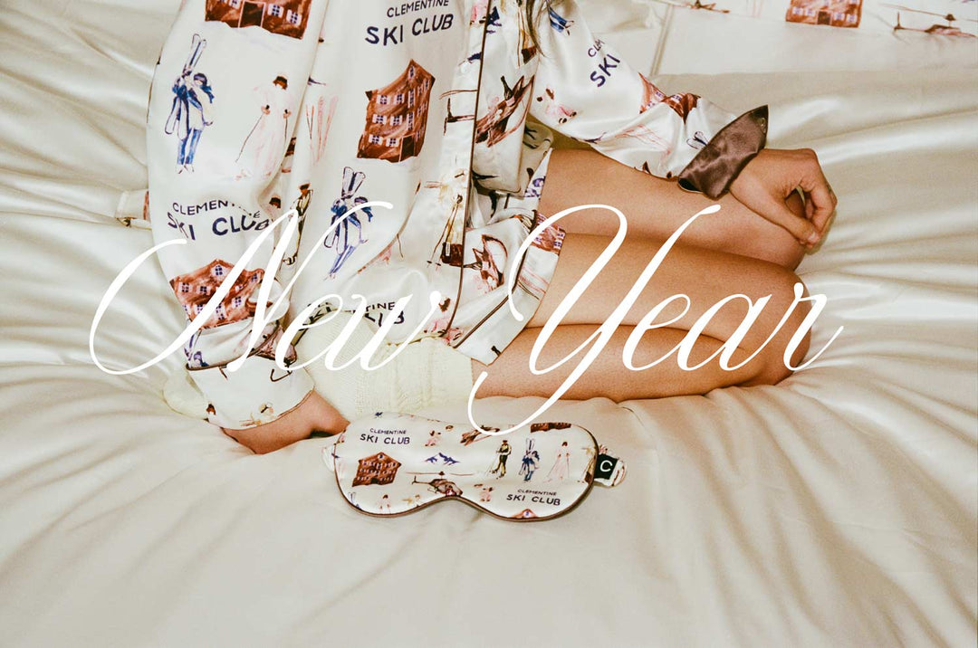 Person seated on bed wearing pajamas and eye mask patterned with CLEMENTINE SKI CLUB; script overlay reads New Year