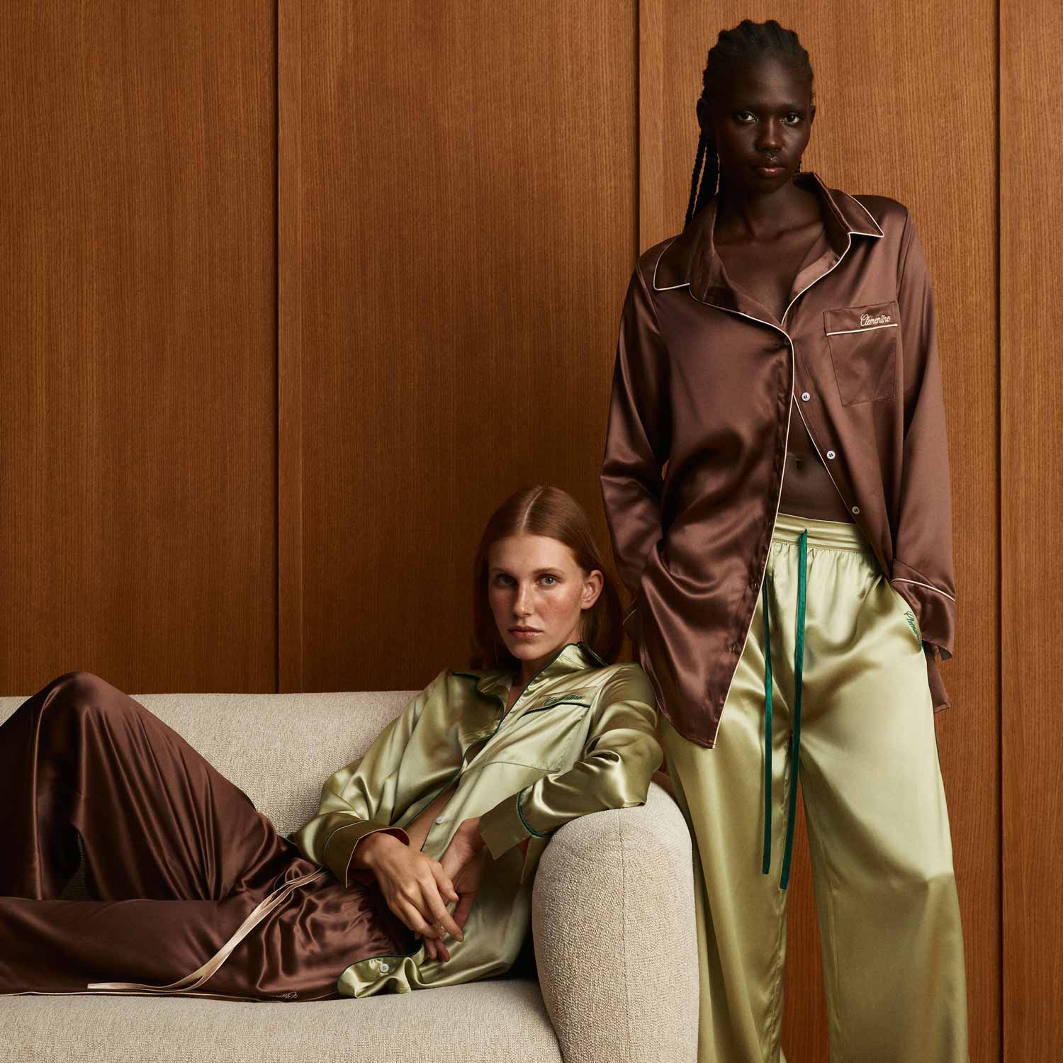 Two models wearing cocoa and sage satin pajama sets against wood panels; standing model's pocket embroidered Clemence. - #color_cocoa