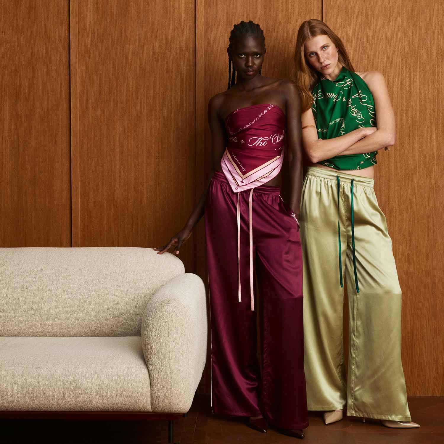 Two women in satin wide-leg pants and scarf-style tops: one in bordeaux strapless set, the other in green top and beige pants, beside a cream sofa. - #color_bordeaux