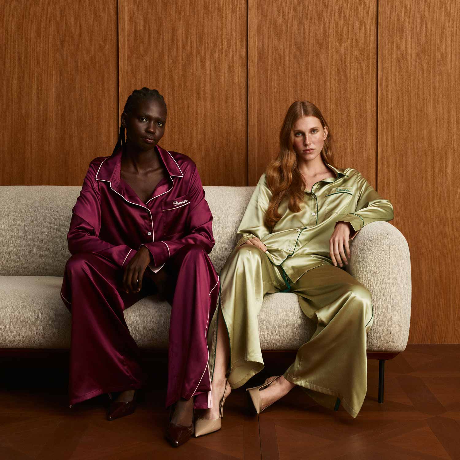 Two women seated on a sofa wearing satin pajama sets: left burgundy, right martini-green, against a wood-paneled wall. - #color_martini-green