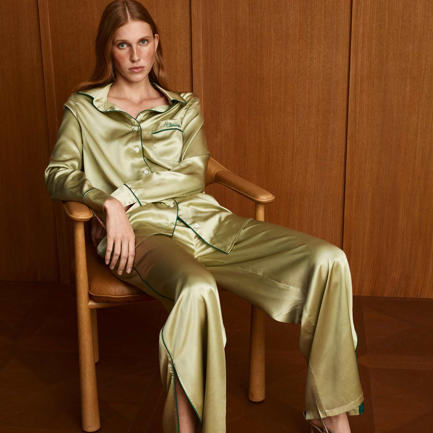 Woman seated in chair wearing martini-green satin pajama set with dark-green piping and 'Clemence' embroidered on chest pocket. - #color_martini-green