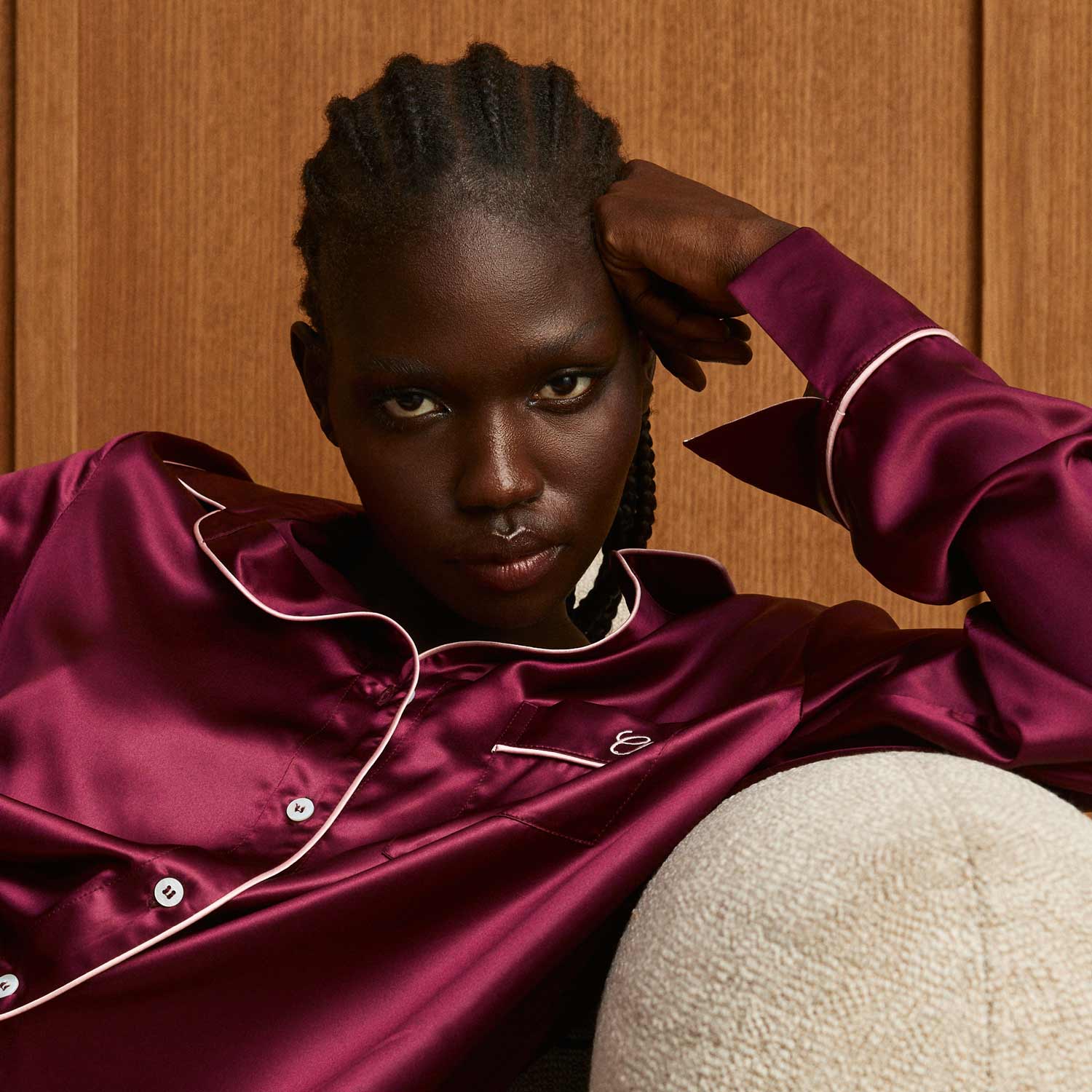 Reclining woman wearing bordeaux silk pajama top with white piping, leaning on a textured cream cushion - #color_bordeaux