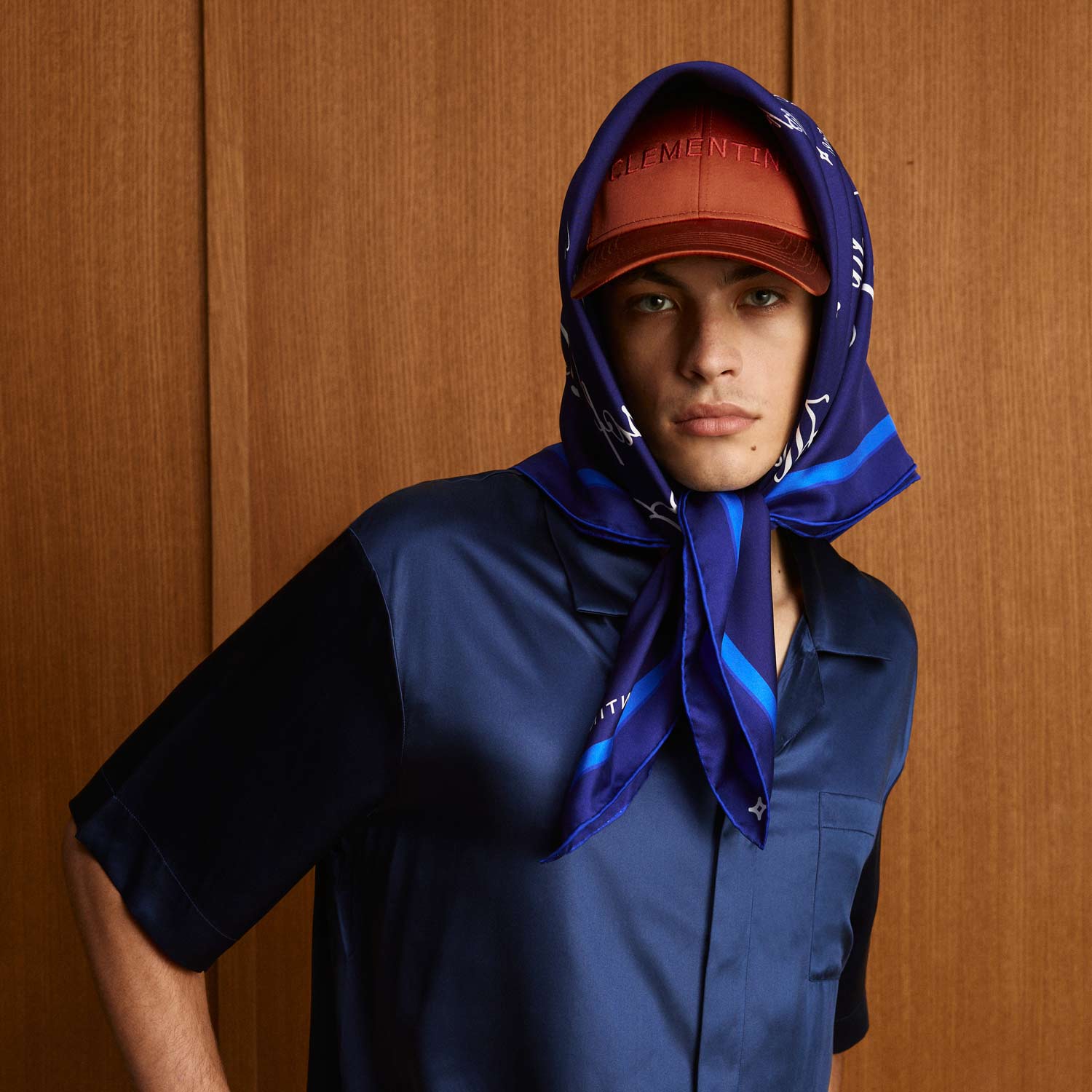Man in navy silk shirt wearing blue patterned headscarf and orange cap embroidered CLEMENTIN against wood background. - #color_midnight-voyage