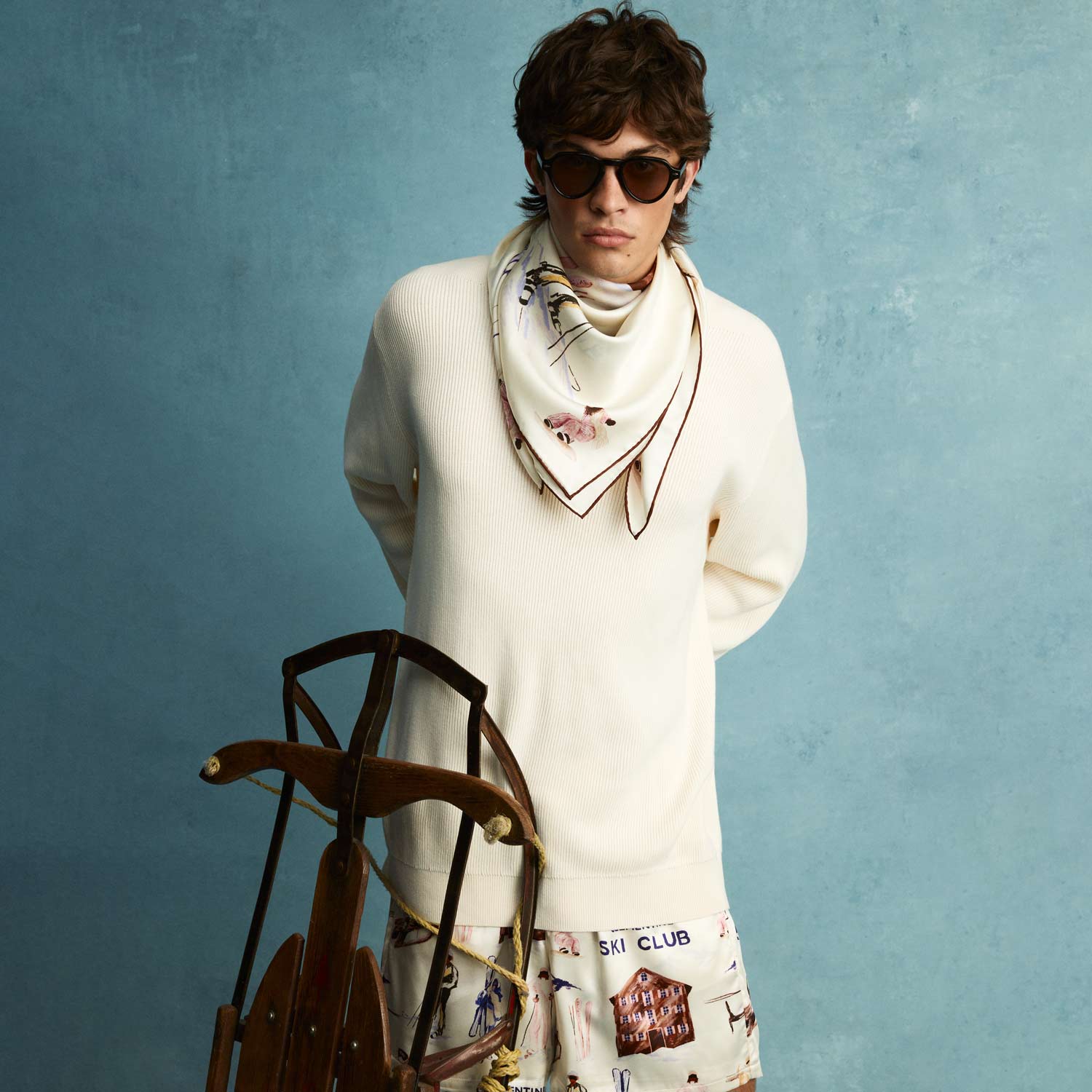 Model wearing cream sweater and floral silk scarf tied at the neck; wooden sled in front; skirt reads SKI CLUB.