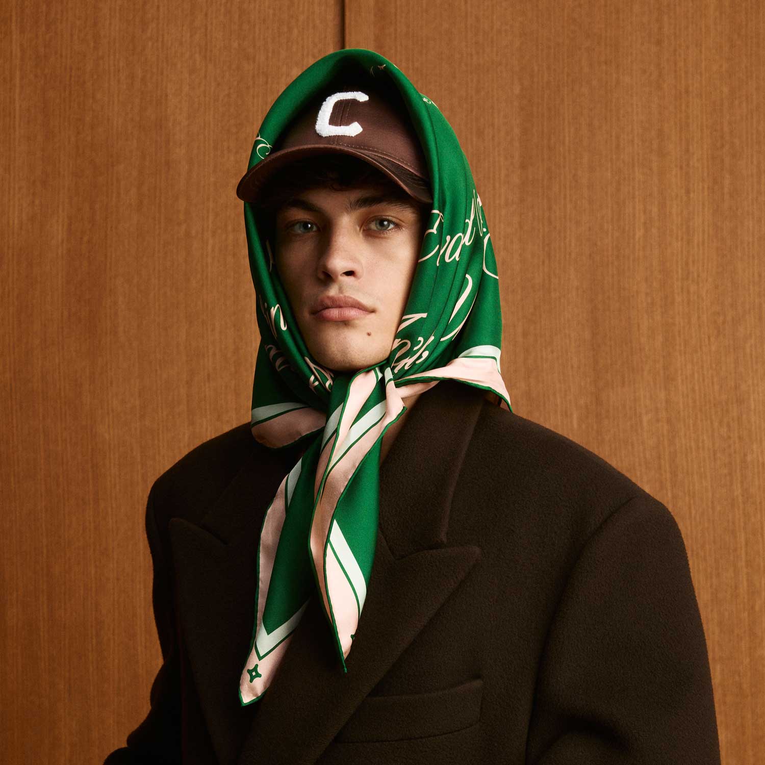 Young man in a dark coat, green silk headscarf tied under chin over a brown cap with a white C, wood-paneled background. - #color_palais-green