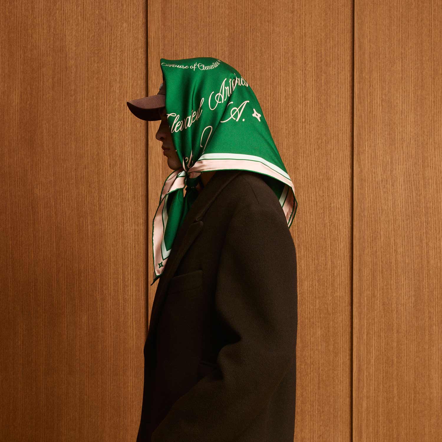 Person in profile wearing green silk scarf tied over a baseball cap; scarf reads 'House of Cleverel Interiors A.'; black coat, wood-paneled background.
