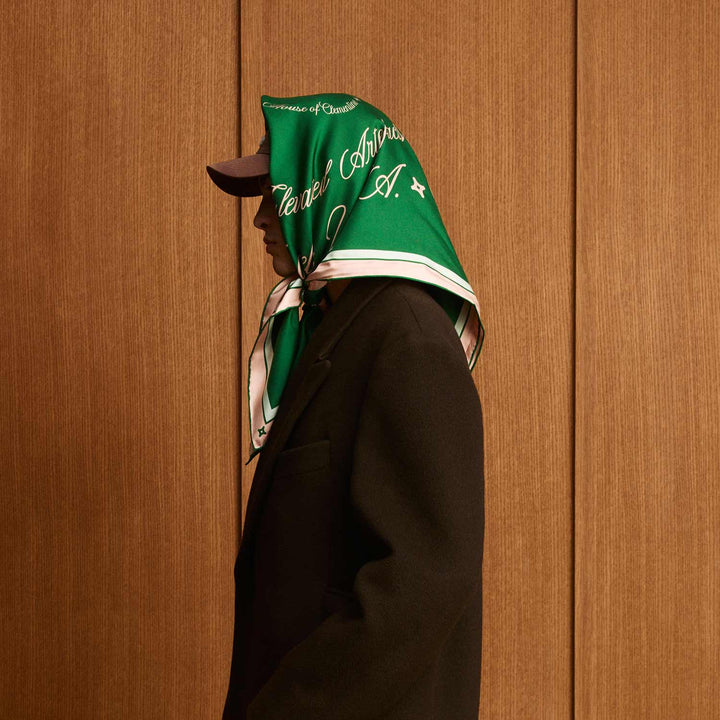 Person in profile wearing green silk scarf tied over a baseball cap; scarf reads 'House of Cleverel Interiors A.'; black coat, wood-paneled background.