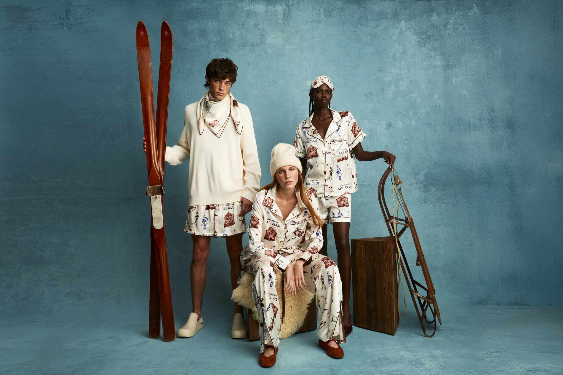 Three models in cream loungewear printed SKI CLUB, one with a beanie, posed with vintage skis and sled on blue background.