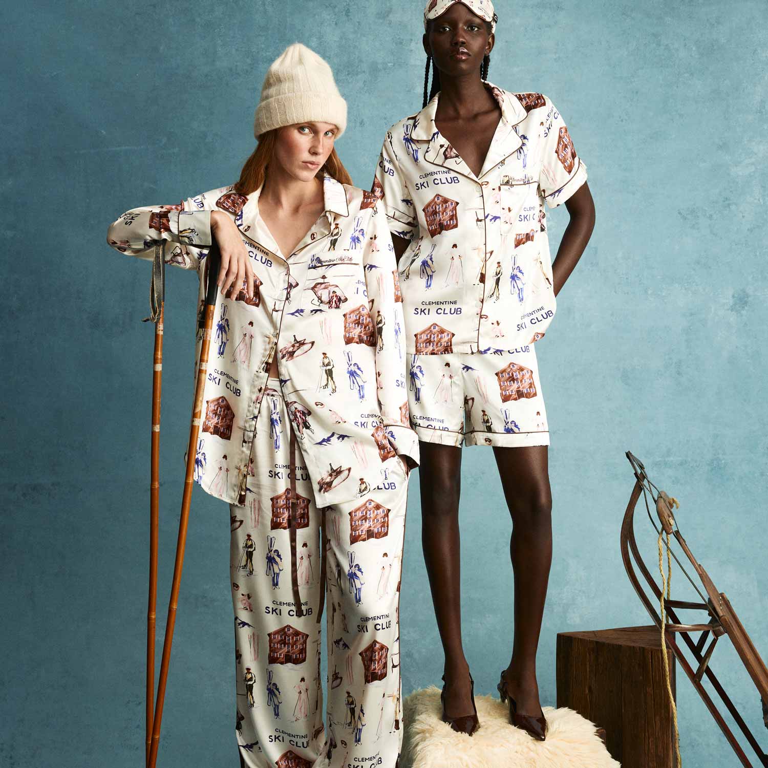 Two models wearing matching 'CLEMENTINE SKI CLUB' pajamas; one leans on ski poles, the other stands on a stool next to a sled.