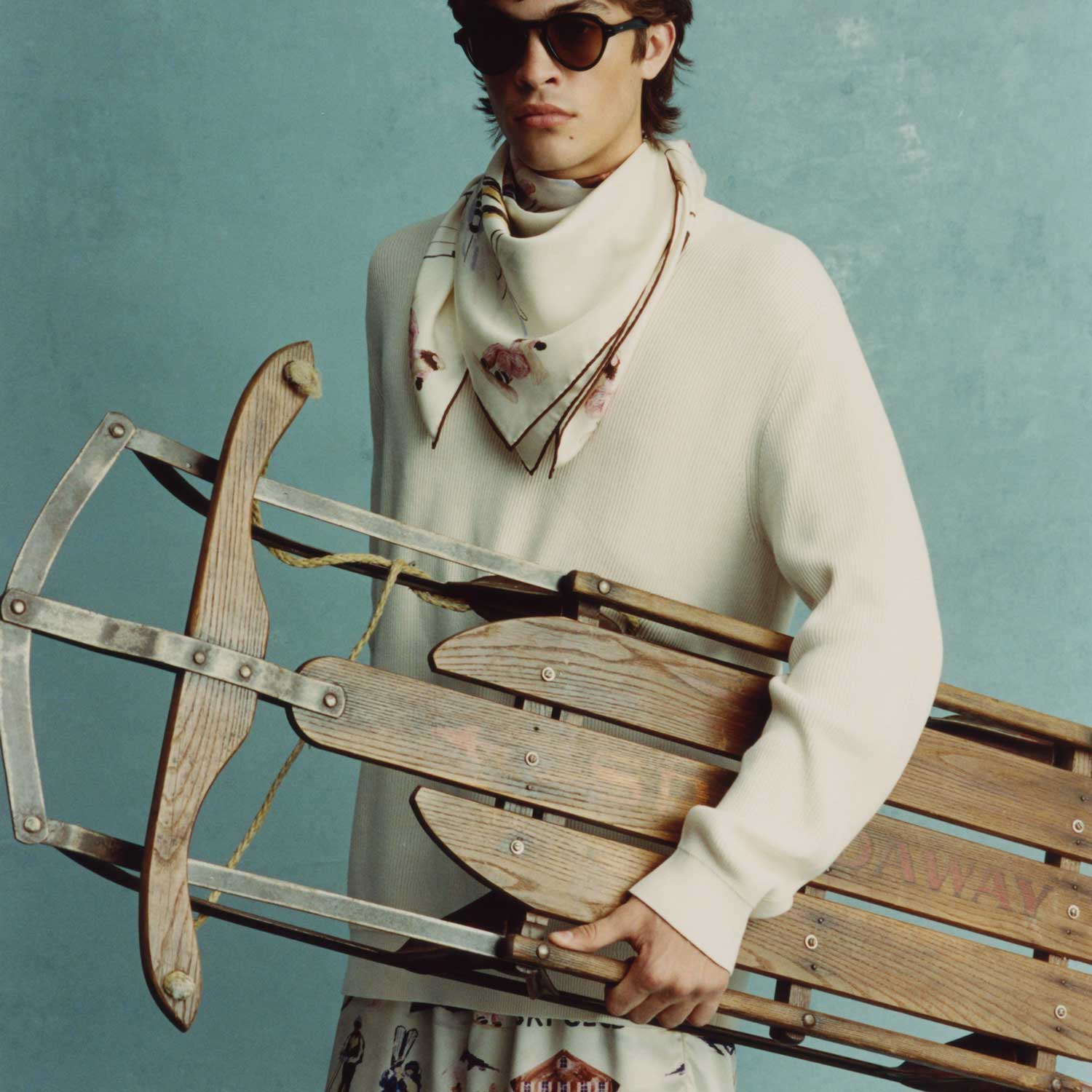 Young man in sunglasses, cream sweater and floral silk scarf holding a vintage wooden sled against a blue backdrop.