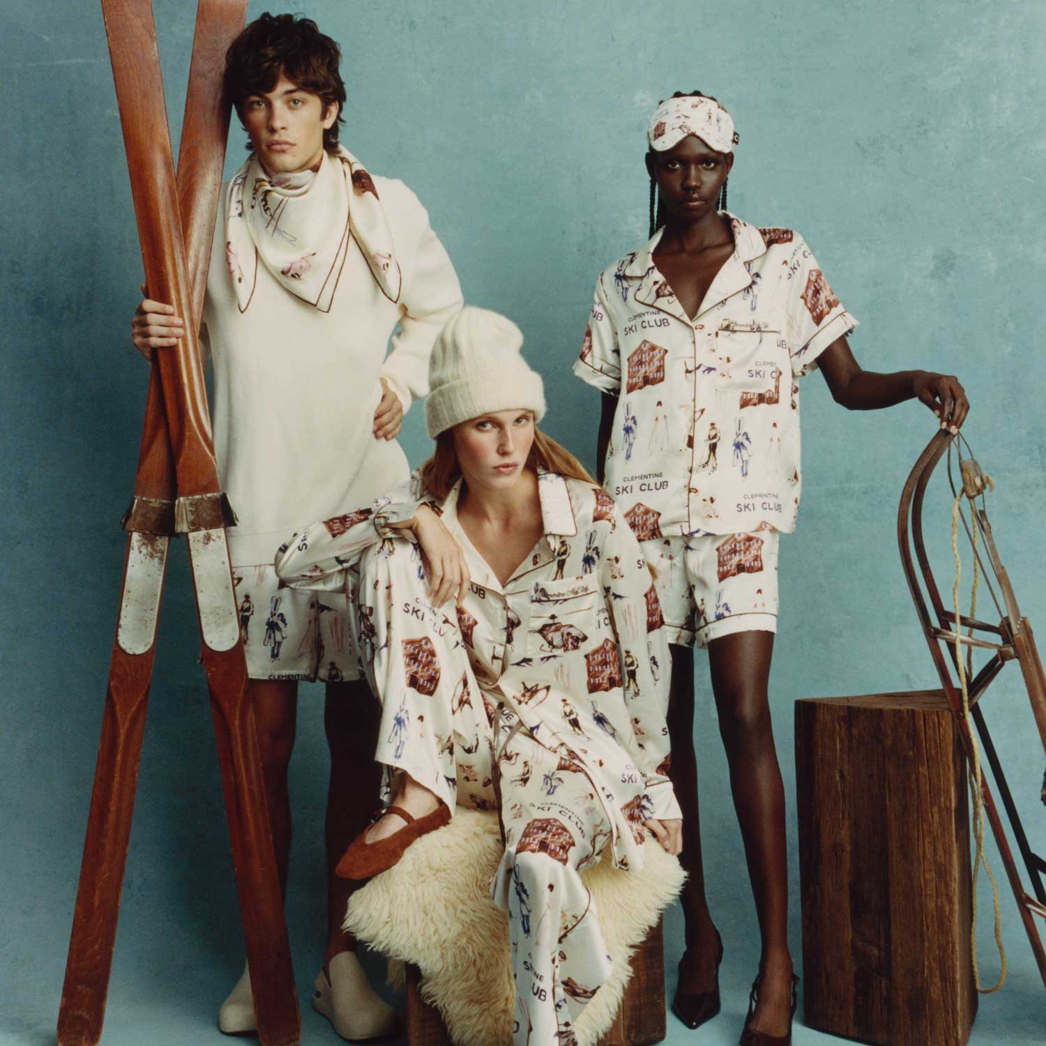 Three models in pajamas printed CLEMENTINE SKI CLUB; center model wears knit beanie; wooden skis and sled on teal backdrop