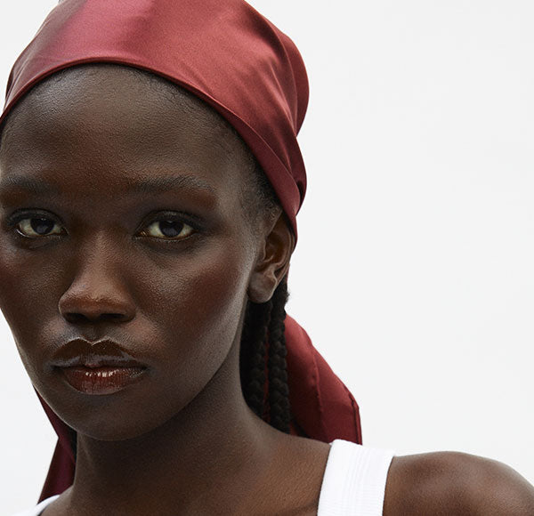 Close-up portrait of a person with braided hair wearing a red headscarf and white tank top, neutral expression.