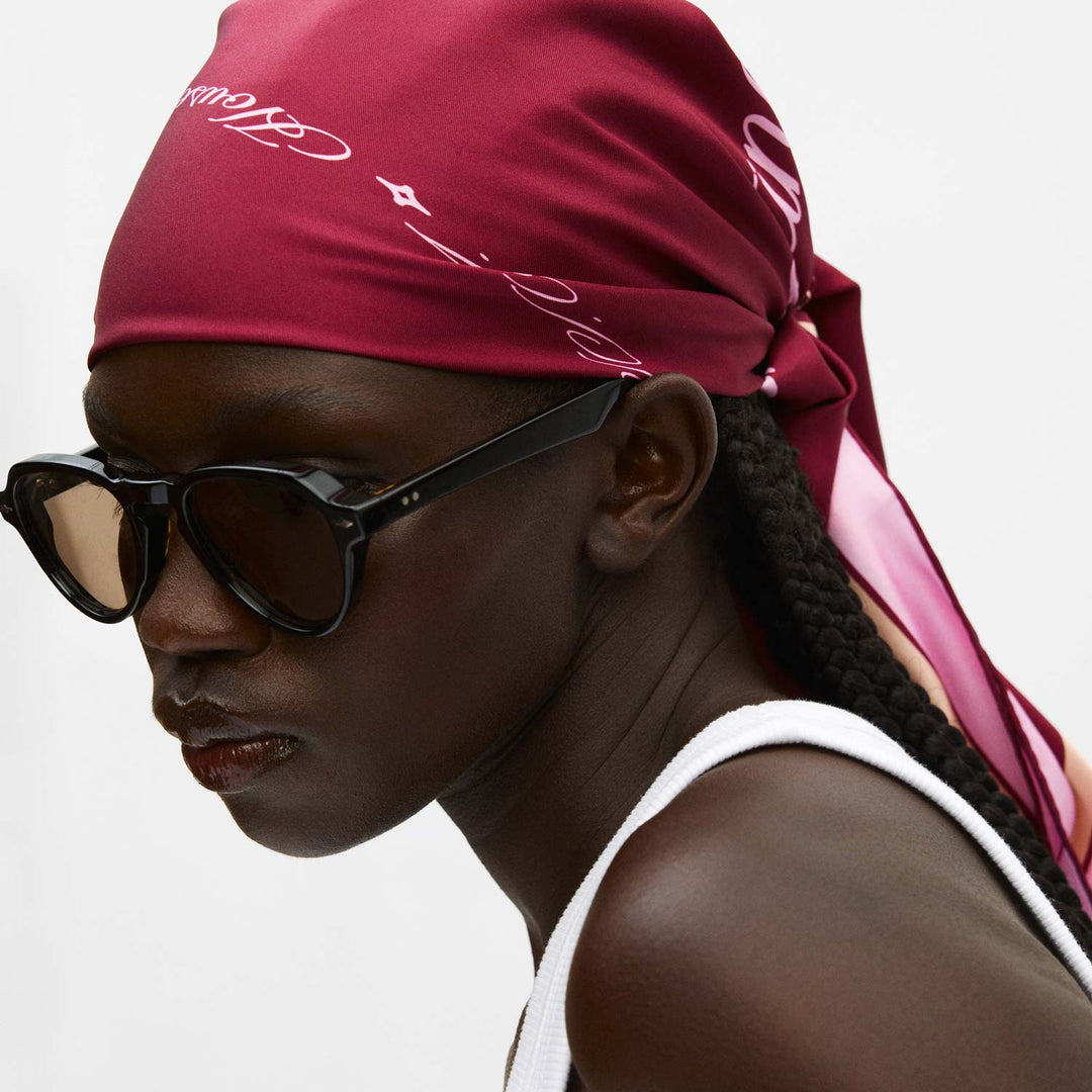 Dark-skinned woman in sunglasses wearing a Mulberry velvet headscarf tied at back, white tank top, braided hair - #color_mulberry-velvet