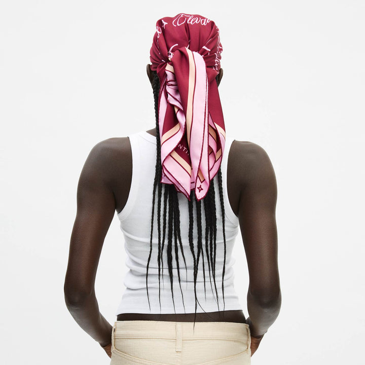 Rear view of model in white tank with long braids, wearing pink-red silk headscarf tied at back; scarf reads 'Elevate'.