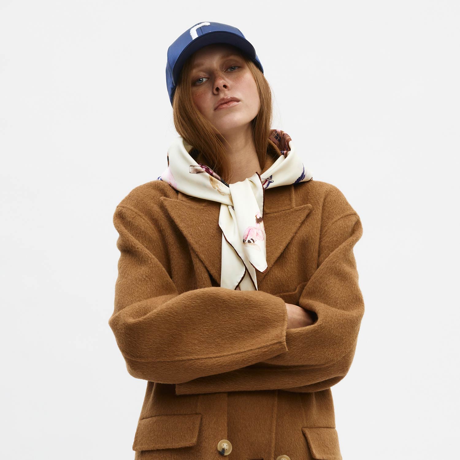 Woman in camel coat with silk scarf tied at neck, arms crossed, wearing blue cap with letter C