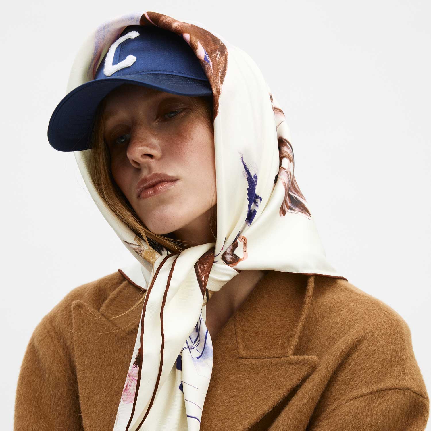Woman wearing cream printed silk headscarf tied under chin, navy cap with letter C, and camel coat.