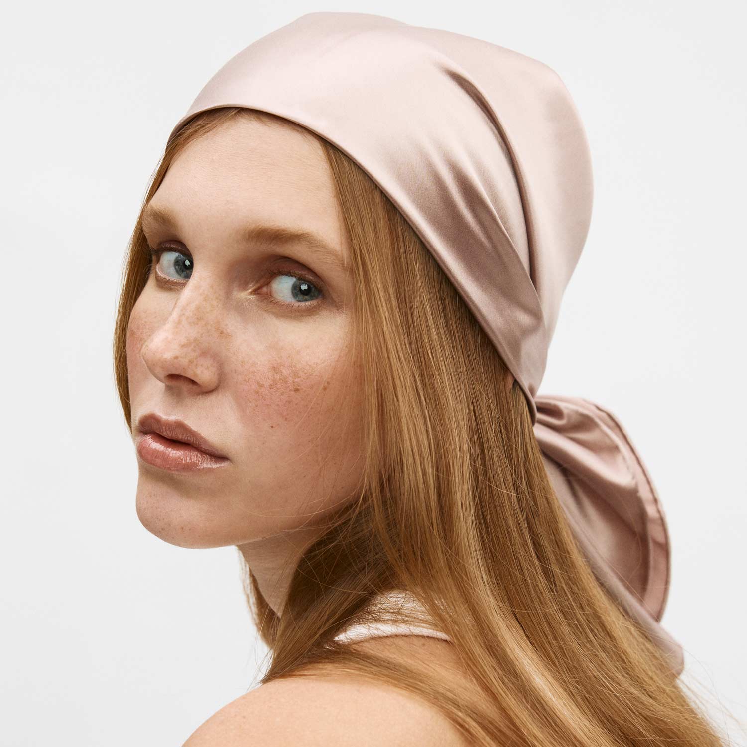 Freckled young woman with long red hair wearing a pale pink satin headscarf, looking over her shoulder.