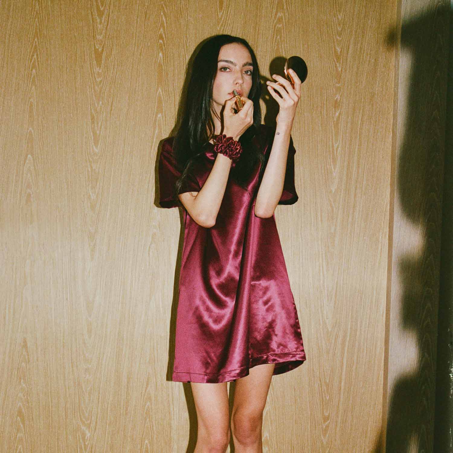 Woman in bordeaux silk mini dress applying lipstick while holding a round compact mirror against a wood-paneled wall #color_bordeaux