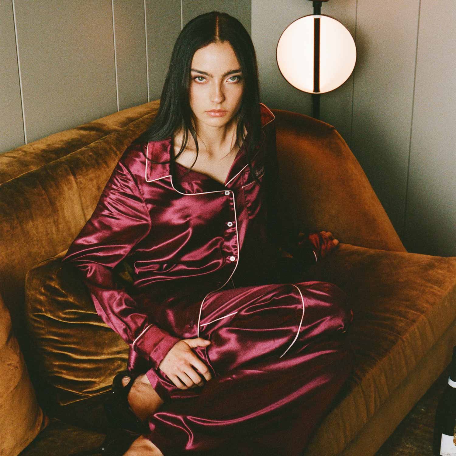 Woman wearing bordeaux silk pajama set sitting on a mustard velvet sofa, gazing at the camera with lamp behind her. #color_bordeaux